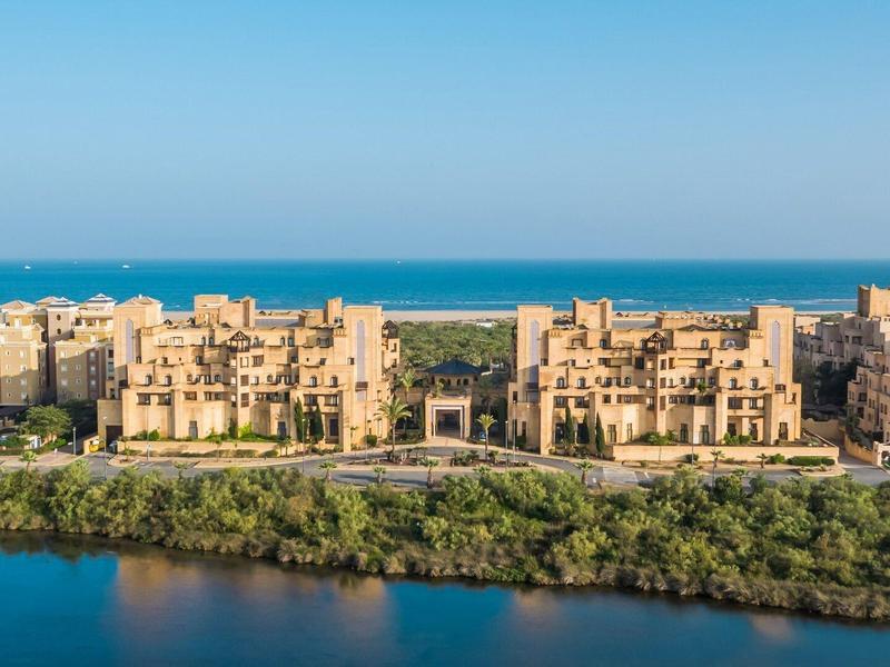 Vista de una ciudad costera con edificios beige, rodeada de agua y vegetación verde.