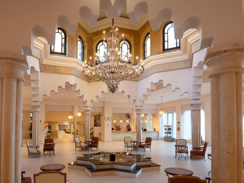 Vestíbulo de hotel iluminado con arcos blancos, columnas y candelabro de estilo marroquí.