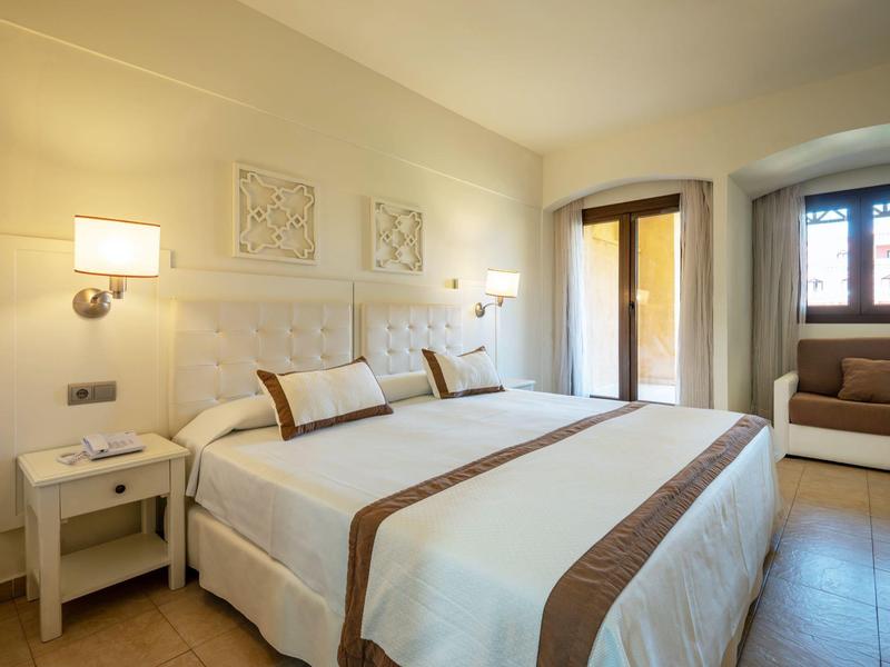 Acogedora habitación doble con paredes claras, dos camas, mesas de noche y sofá junto a la ventana.