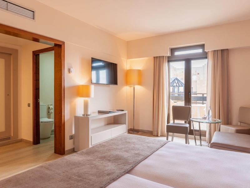 Habitación de hotel luminosa con dos camas, área de estar y balcón con vista al exterior.