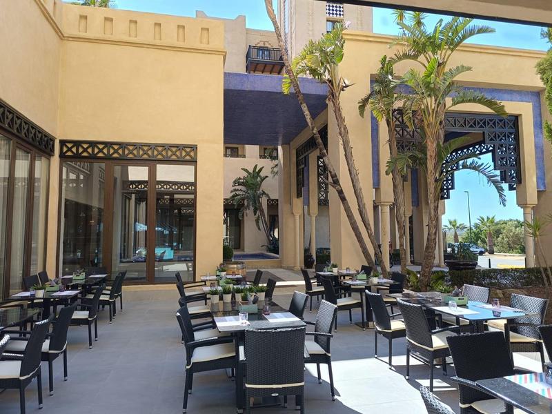 Patio al aire libre de un hotel con mesas y sillas negras rodeado de arquitectura tradicional y palmeras.