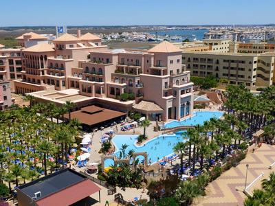 Grand complexe avec plusieurs bâtiments et une grande piscine entourée de palmiers.