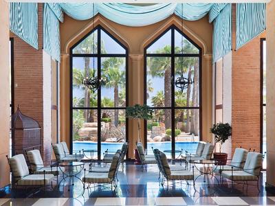 Grande salle à manger lumineuse avec hautes fenêtres et vue sur des palmiers.