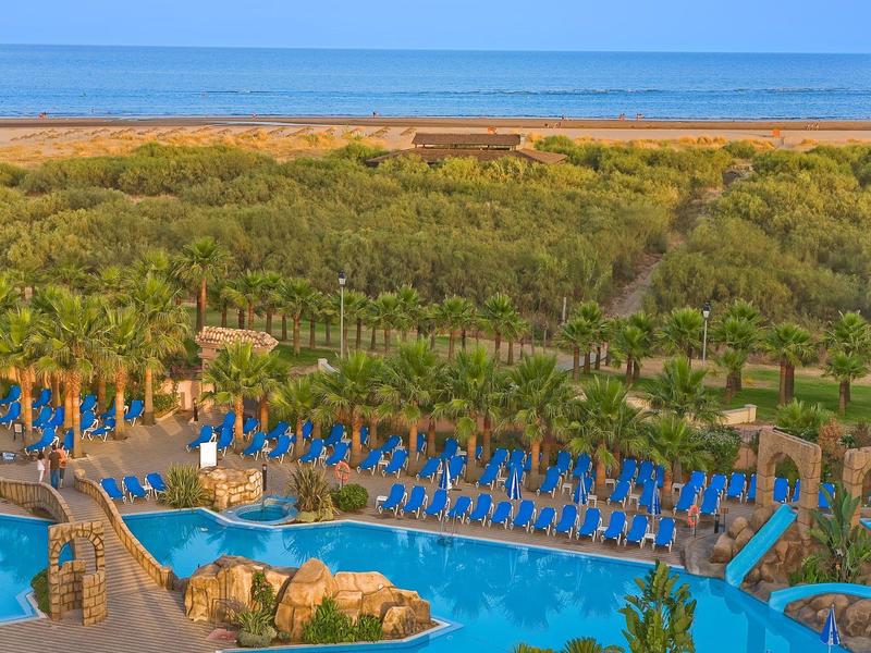 Piscine d'hôtel entourée de parasols bleus, vue sur la plage et la mer au loin.