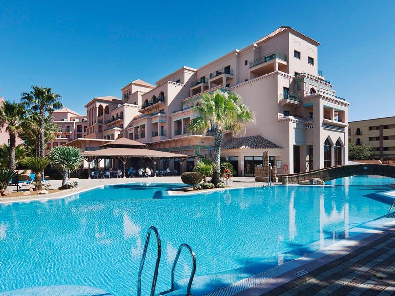 Hôtel avec grande piscine, palmiers et ciel ensoleillé pour des vacances relaxantes.