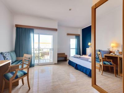 Helles Hotelzimmer mit Doppelbett, Schreibtisch, Stühlen, blau-weißer Farbgestaltung und Balkon.