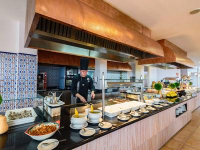 Buffet mit vielfältigen Speisen, Küchenchef hinter der Theke, helle Fliesen und moderne Holztheke.