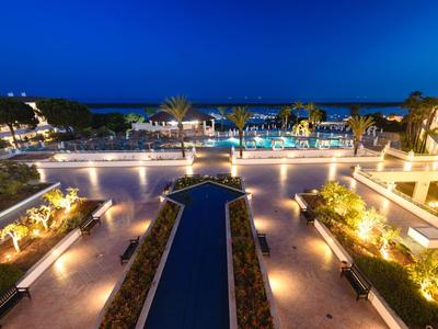 Luxurious hotel with illuminated pool and gardens at night, sea in the background.