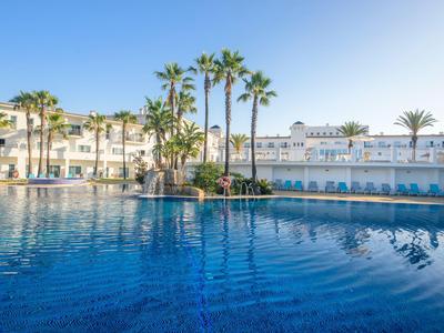 Large outdoor pool with palm trees and hotel building in the background under clear sky.