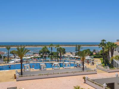 Blick auf einen Poolbereich mit Liegestühlen, Palmen und dahinter das blaue Meer unter klarem Himmel.