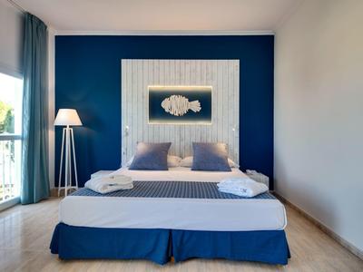 Schlafzimmer mit großem Bett, blau-weißer Wand, zwei Kissen, Stehlampe und Balkonfenster.