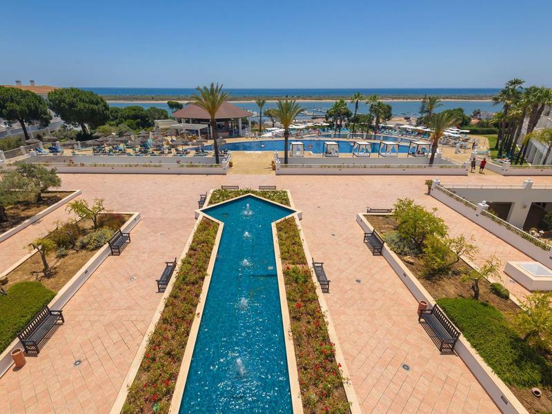 Großer Pool mit Pfeilform, Palmen, Sitzbereichen und Meer im Hintergrund an sonnigem Tag.