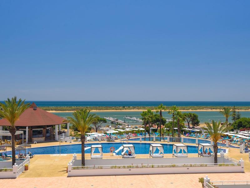 Großer Pool mit Liegestühlen und Palmen, dahinter Sandstrand und ruhiges Meer unter blauem Himmel.