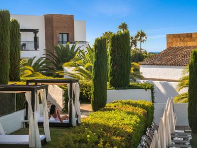 Modernes Hotel mit grünen Gärten, hohen Zypressen und weißen Sonnenterrassen unter blauem Himmel.