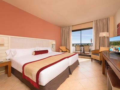 Camera d'hotel luminosa con letto grande, scrivania, TV e balcone con vista sul mare.