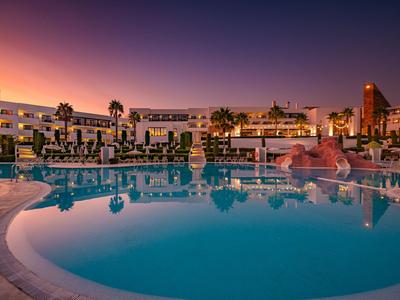 Hotel mit großem Außenpool bei Sonnenuntergang mit Palmen und Liegestühlen