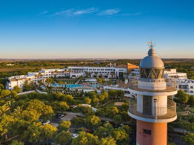 Faro davanti a un grande edificio alberghiero con piscine, circondato da paesaggio verde sotto un cielo azzurro.