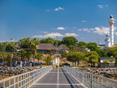 Blick auf einen Pier mit Palmen und Leuchtturm am Ende unter blauem Himmel.