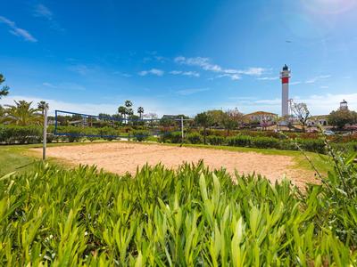 Una spiaggia soleggiata con una rete da pallavolo e un faro sullo sfondo, circondata da piante.