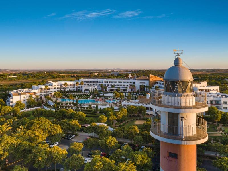 Faro davanti a un grande edificio alberghiero con piscine, circondato da paesaggio verde sotto un cielo azzurro.