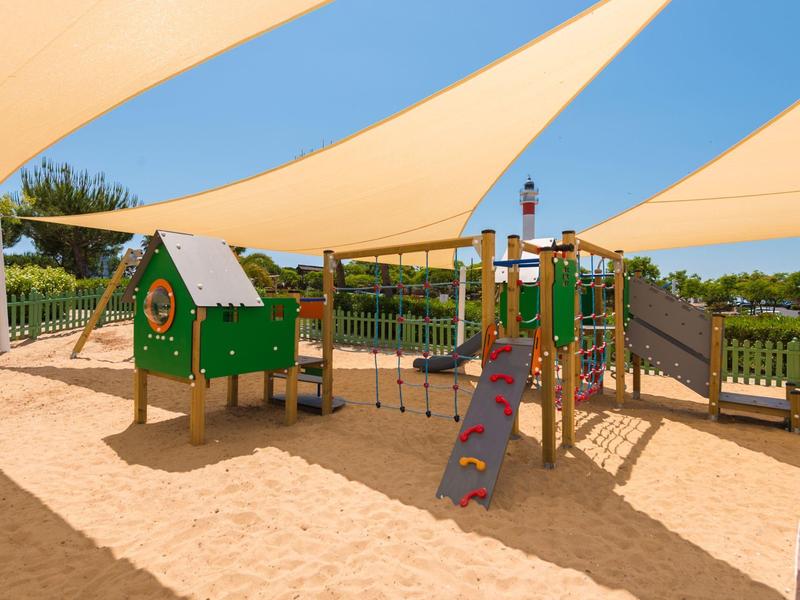 Un parco giochi ombreggiato con sabbia, rete da arrampicata e scivolo sotto un cielo sereno.