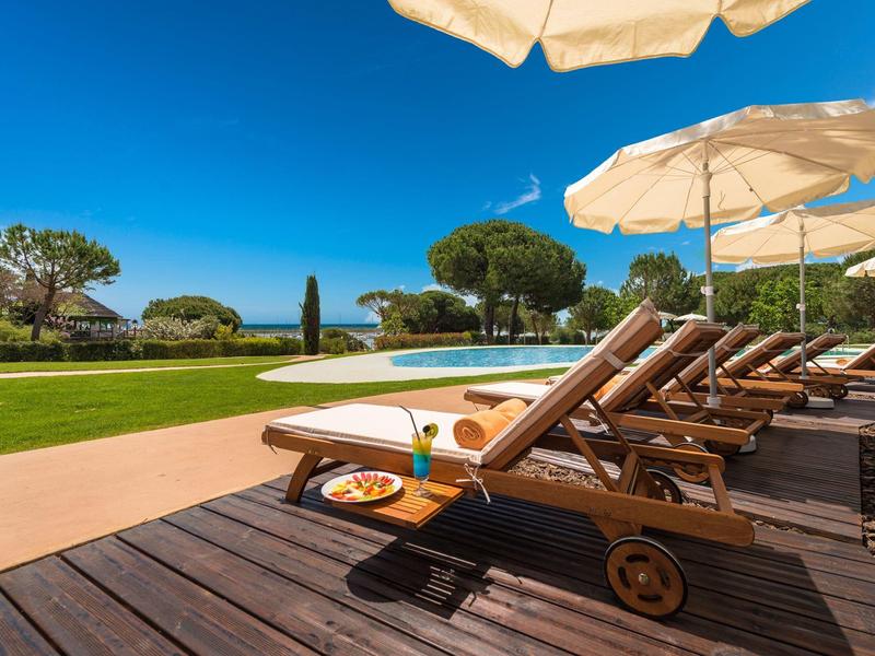 Lettini a bordo piscina con ombrelloni, vista sul giardino e sul mare sotto un cielo azzurro.