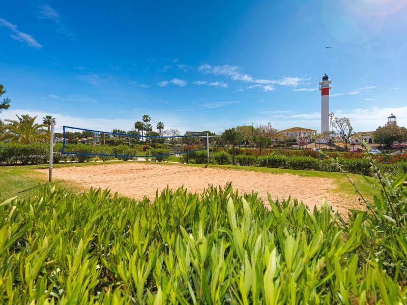 Una spiaggia soleggiata con una rete da pallavolo e un faro sullo sfondo, circondata da piante.