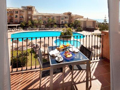 Balkon mit Tisch und zwei Stühlen, Pool und Hotelgebäude im Hintergrund bei Sonnenschein.