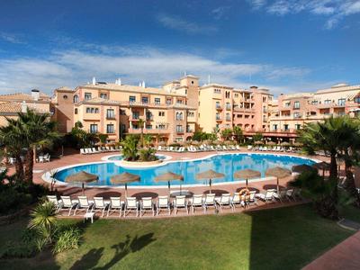 Hotelcomplex met een groot rond zwembad omringd door ligstoelen en palmbomen.