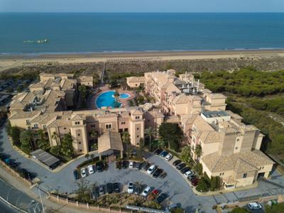 Een kusthotelcomplex met een bochtige oprijlaan, parkeerplaats, zwembad en een nabijgelegen strand.
