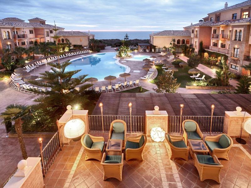 Luxe resort met groot zwembad, ligbedden, palmbomen en twee verdiepingen tellende gebouwen onder bewolkte lucht.