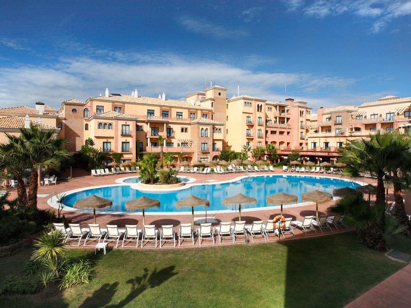 Hotelcomplex met een groot rond zwembad omringd door ligstoelen en palmbomen.