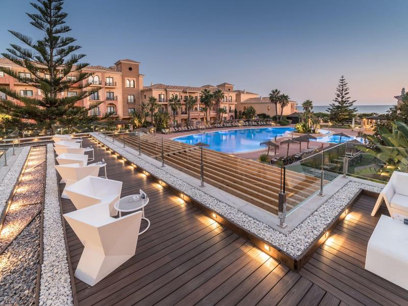 Luxe resort met verlichte houten vlonder, witte stoelen, zwembad en hotelgebouwen bij zonsondergang.