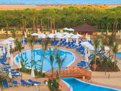 Großer blauer Swimmingpool mit Brücke, Liegestühlen und grünen Palmen nahe dem Meer.