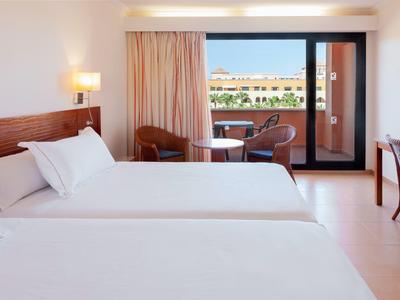 Hell eingerichtetes Hotelzimmer mit zwei großen Betten, Balkon und Holzmöbeln in warmen Farbtönen.