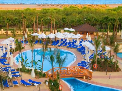 Hotelpoolanlage mit runden Pools, blauen Liegestühlen, Holzbrücke und Blick auf das Meer.