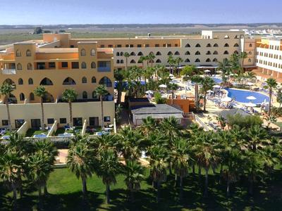 Großer Hotelkomplex mit Pool, Garten und Palmen unter blauem Himmel an Küste.