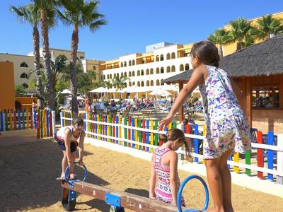 Kinder spielen im Sand unter Palmen vor bunten Zäunen und gelben Gebäuden bei Sonnenschein.