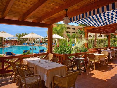 Überdachte Terrasse mit Tischen und Stühlen, Blick auf Pool, Palmen und sonnigen Himmel.