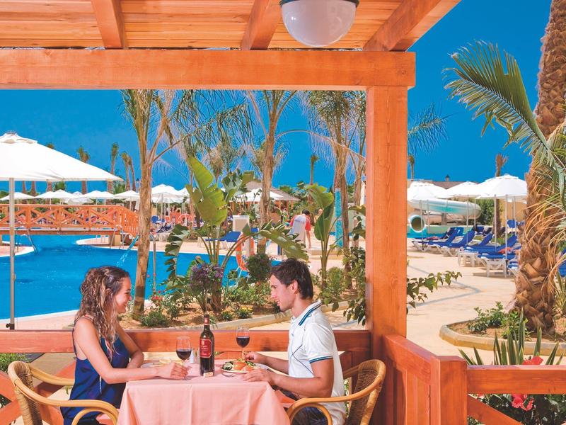 Paar sitzt an gedecktem Tisch unter Holzpergola, im Hintergrund Pool, Sonnenschirme und Palmen.