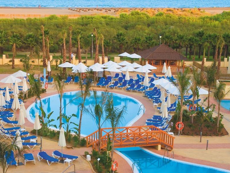 Großer blauer Swimmingpool mit Brücke, Liegestühlen und grünen Palmen nahe dem Meer.