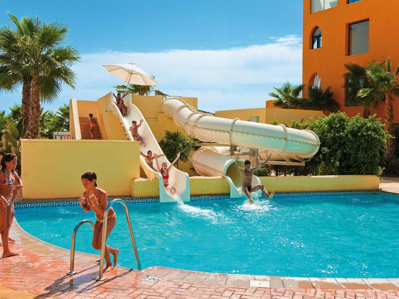 Menschen spielen und rutschen an einem sonnigen Tag im blauen Pool neben einem orangen Gebäude.