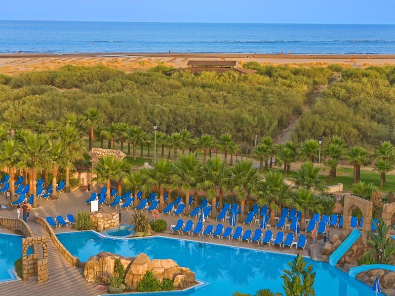 Großer Pool mit blauen Liegestühlen, umgeben von grüner Vegetation und Blick aufs Meer im Hintergrund.