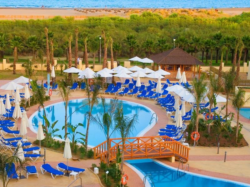 Hotelpoolanlage mit runden Pools, blauen Liegestühlen, Holzbrücke und Blick auf das Meer.