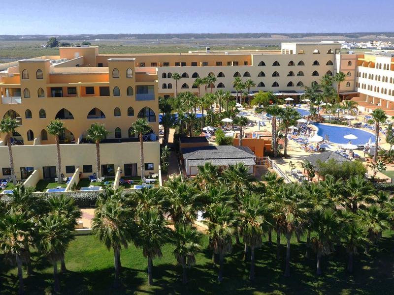 Großer Hotelkomplex mit Pool, Garten und Palmen unter blauem Himmel an Küste.