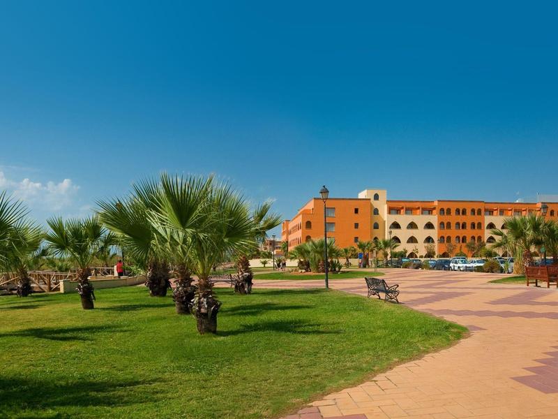 Grüner Park mit Palmen, breitem gepflastertem Weg und mehrstöckigem orange-gelben Gebäude im Hintergrund.