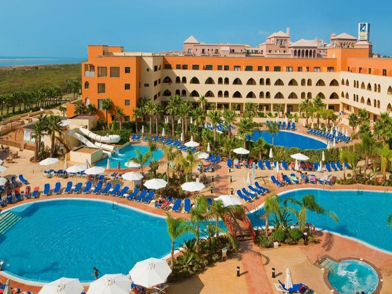 Großes Hotel mit drei blauen Pools, Palmen, Sonnenschirmen und weitem Blick auf das Meer.