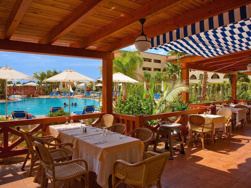 Überdachte Terrasse mit Tischen und Stühlen, Blick auf Pool, Palmen und sonnigen Himmel.