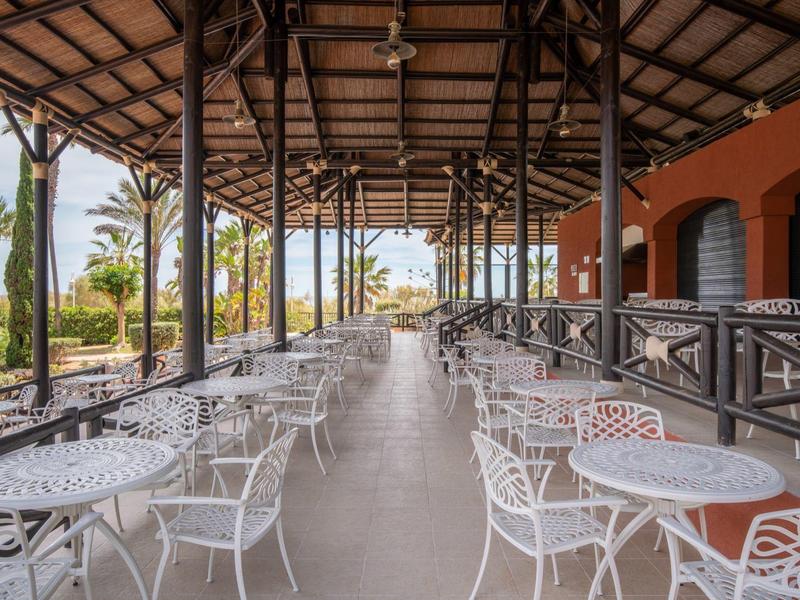 Überdachte Terrasse mit weißen Metalltischen und Stühlen, Boden aus grauem Beton, Palmen im Hintergrund.