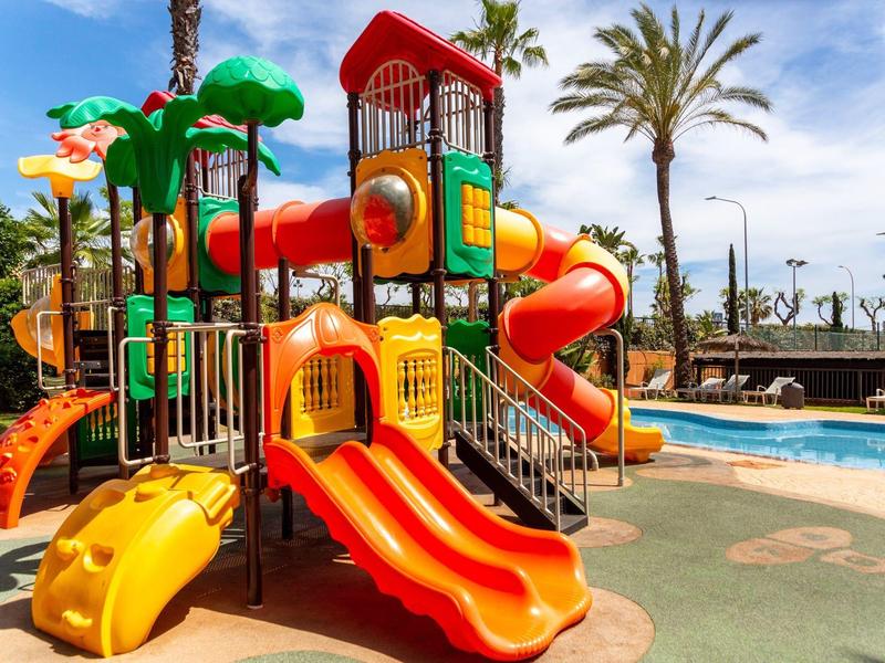 Bunter Spielplatz mit Rutschen und Klettergerüsten neben einem Schwimmbecken unter blauem Himmel.